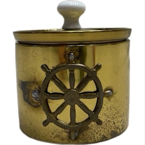 May include: A cylindrical brass container with a lid featuring a white ceramic knob. The front of the container is decorated with a raised brass ship's wheel emblem. The brass surface shows signs of wear and patina, with visible metallic texture.