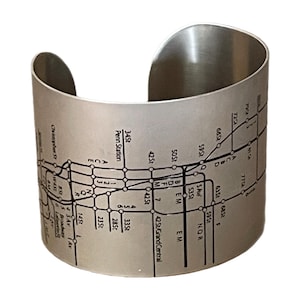 May include: A silver-toned metal cuff bracelet featuring a black engraved map of the New York City subway system. The design includes various station names such as Penn Station and 42 St/Grand Central, along with line identifiers like A, C, E, 1, 2, 3, 7, B, D, F, M, N, Q, R, and L.