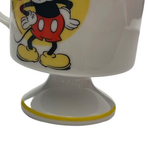 May include: A white ceramic pedestal mug featuring a graphic of Mickey Mouse standing against a yellow circle. The character wears red shorts and yellow shoes. The text Walt Disney Productions is printed vertically in black near the handle.