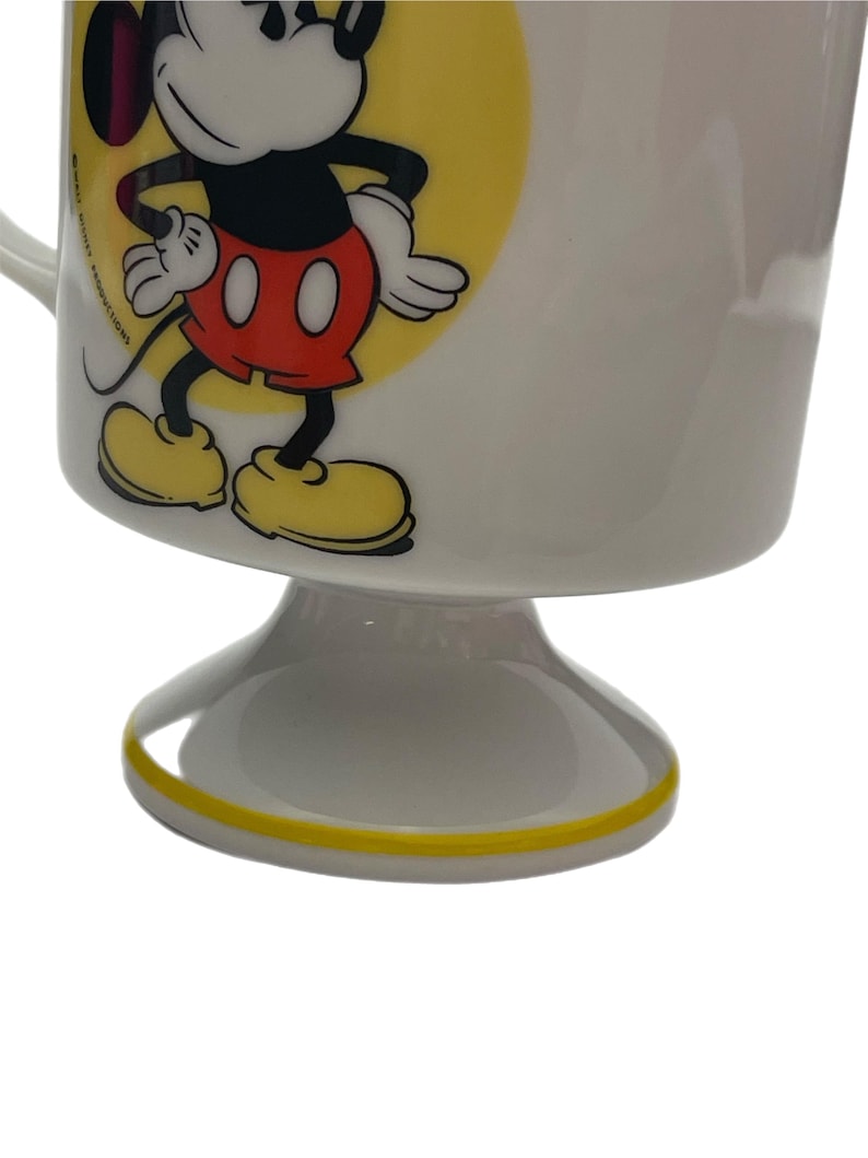 May include: A white ceramic pedestal mug featuring a graphic of Mickey Mouse standing against a yellow circle. The character wears red shorts and yellow shoes. The text Walt Disney Productions is printed vertically in black near the handle.