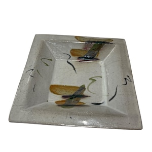 May include: Square ceramic dish with a speckled, off-white finish. The dish features abstract brushstrokes in shades of green, yellow, and brown. Black lines and accents add detail to the design. The dish has a slightly recessed center.