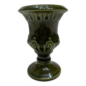 May include: A dark olive green ceramic vase with a glossy finish. The vase features a wide, flared rim with a decorative scalloped edge, a tapered body with vertical embossed detailing, and a sturdy, circular pedestal base for stability.