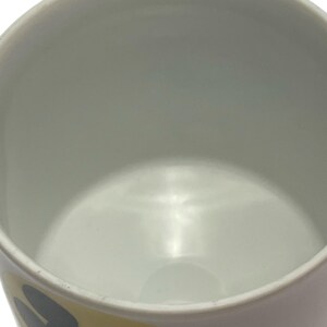 May include: A white ceramic mug featuring a yellow graphic panel on the exterior. The panel displays a black and white illustration of Mickey Mouse. The interior of the mug is plain white, and the rim is smooth and circular, showing a simple design.