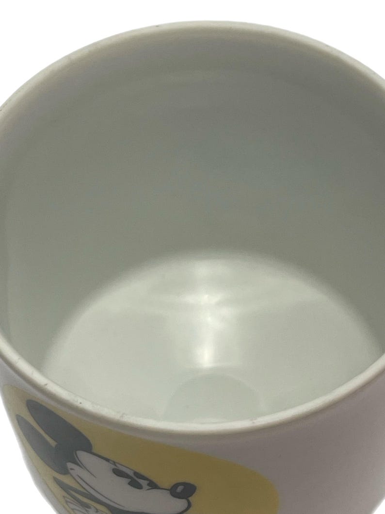 May include: A white ceramic mug featuring a yellow graphic panel on the exterior. The panel displays a black and white illustration of Mickey Mouse. The interior of the mug is plain white, and the rim is smooth and circular, showing a simple design.