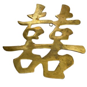 May include: A flat, brass-colored metal wall hanging featuring the Chinese double happiness character. The object has a textured, aged surface with a small metal loop at the top center for hanging. The design consists of symmetrical, stylized strokes.