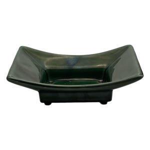 May include: A square, dark green ceramic dish with a glossy finish, featuring slightly curved, flared edges and a raised base supported by small feet. The interior of the bowl is smooth and deep, showcasing a uniform, dark forest green glaze.