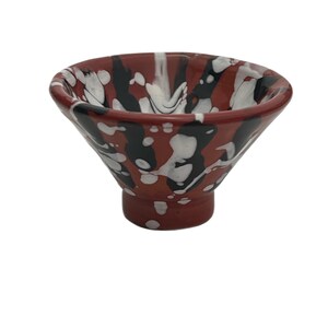 May include: A small, flared ceramic bowl featuring a glossy finish with a marbled pattern of deep red, black, and white colors. The bowl has a wide, open top that tapers down to a narrow, cylindrical base, creating a distinct funnel-like shape.