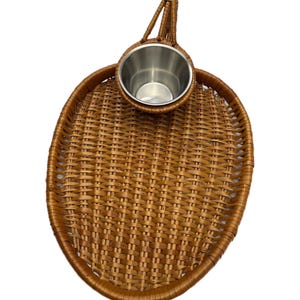 May include: An oval-shaped brown woven wicker tray featuring a single integrated handle at the top and a circular silver-colored metal cup holder insert positioned near the handle, creating a functional serving piece with a natural texture.