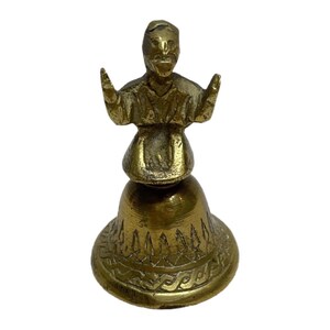 May include: A brass bell featuring a handle shaped like a figure with arms raised. The bell has a flared base decorated with engraved geometric patterns and a series of vertical leaf-like motifs around the lower section of the metal body.
