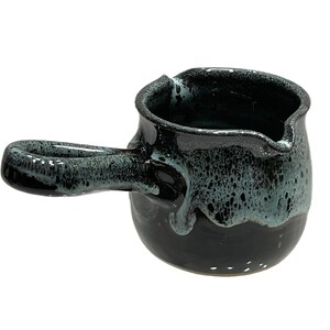May include: A small, black ceramic creamer with a green glaze and a handle. The creamer has a speckled, uneven glaze.