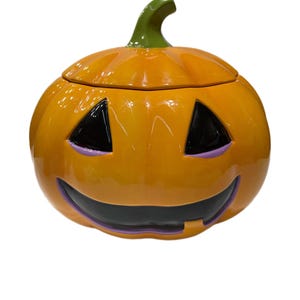 May include: A ceramic pumpkin-shaped container with a lid, perfect for Halloween. The pumpkin is orange with a green stem and features a carved jack-o'-lantern face with black eyes and mouth, and purple accents. The container is glossy.