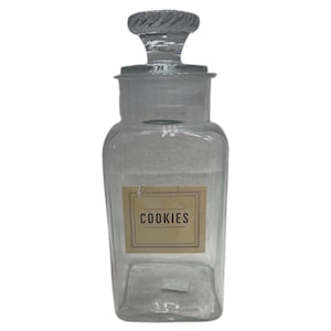 May include: A clear glass jar with a square base and a matching glass lid featuring a twisted knob handle. A rectangular beige label is centered on the front of the jar, displaying the word COOKIES in black capital letters within a thin border.