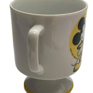 May include: A white ceramic mug with a pedestal base and a yellow rim accent. The side features a graphic of Mickey Mouse against a yellow circular background, with the text Walt Disney Productions printed in small black letters near the character.