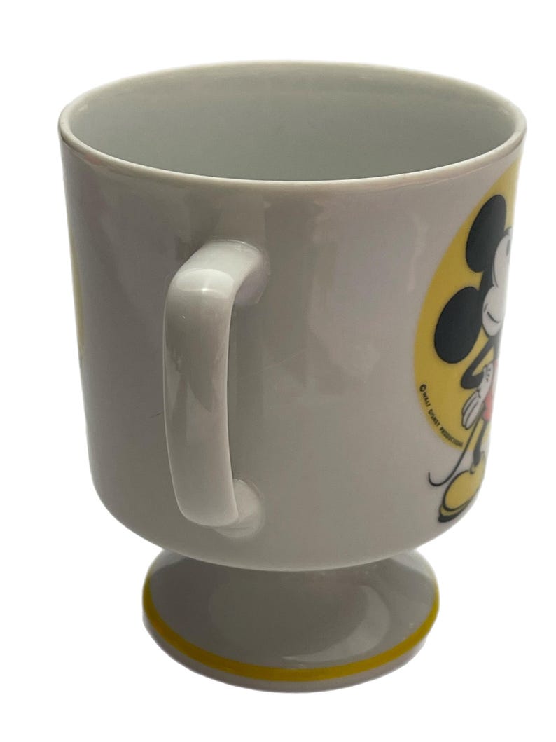 May include: A white ceramic mug with a pedestal base and a yellow rim accent. The side features a graphic of Mickey Mouse against a yellow circular background, with the text Walt Disney Productions printed in small black letters near the character.