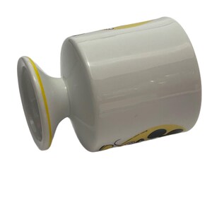 May include: A white ceramic mug with a pedestal base featuring a yellow rim and a graphic design on the side. The mug has a smooth, glossy finish and a cylindrical body that tapers slightly toward the base, showcasing a yellow and black pattern.
