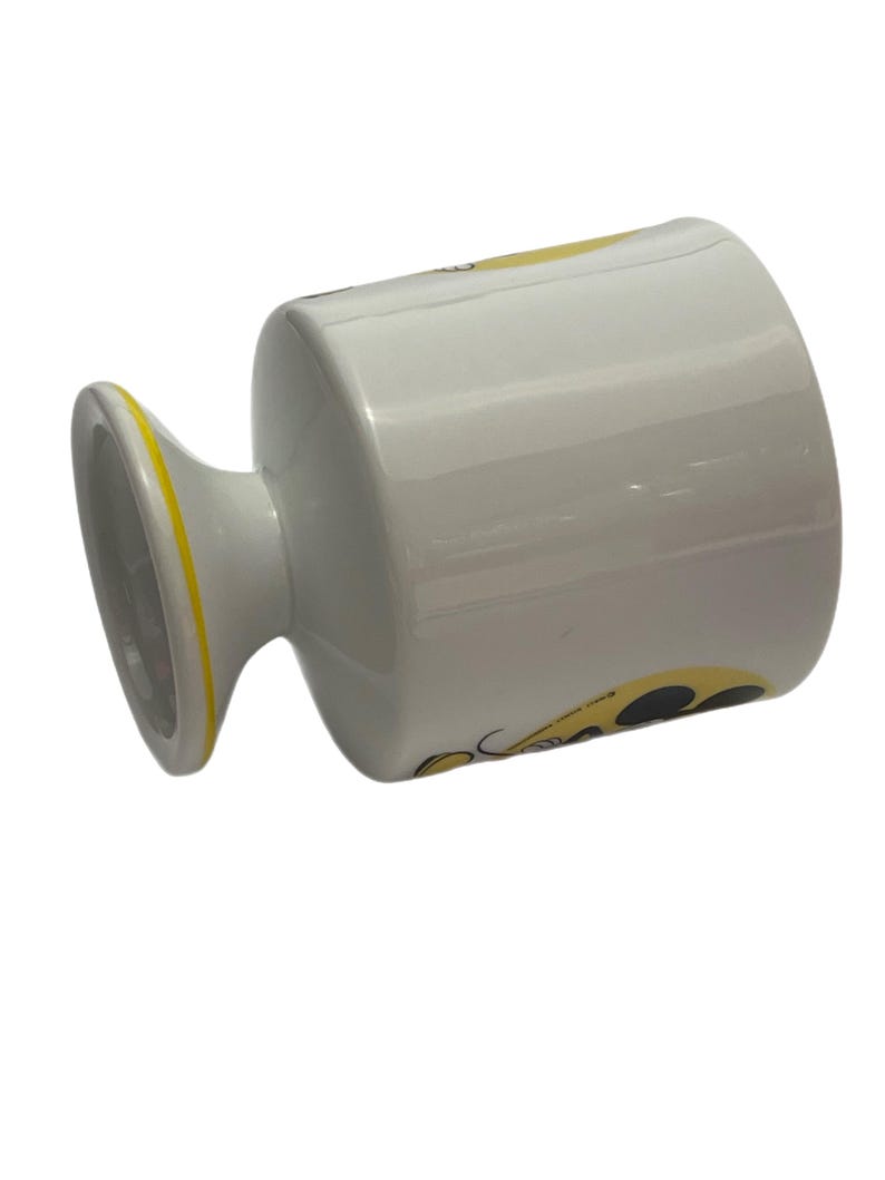 May include: A white ceramic mug with a pedestal base featuring a yellow rim and a graphic design on the side. The mug has a smooth, glossy finish and a cylindrical body that tapers slightly toward the base, showcasing a yellow and black pattern.