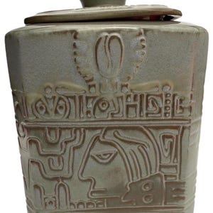 May include: A square ceramic canister with a lid, featuring a light green glaze and embossed relief designs. The front displays a stylized profile of a face and geometric patterns, with a coffee bean symbol centered near the top rim.