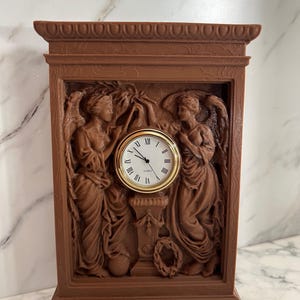 Può includere: Un orologio in legno marrone con un quadrante dell'orologio con numeri romani e due figure su entrambi i lati. L'orologio è in una cornice decorativa con un design in rilievo.