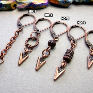 Puede incluir: Cinco pendientes de flecha de cobre con diferentes estilos. Los colgantes están unidos a un pendiente de aro con cierre de gancho. Los estilos incluyen una cadena, un alambre retorcido, una piedra preciosa roja, un cilindro texturizado y una punta de flecha simple.