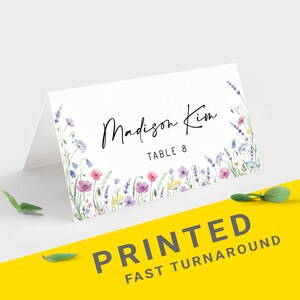 Pu&ograve; includere: Un segnaposto bianco piegato con un design floreale ad acquerello. La carta presenta il nome "Madison Kim" e "Table 8" in corsivo nero. L'angolo in basso a destra riporta il testo "PRINTED FAST TURNAROUND" in grigio.