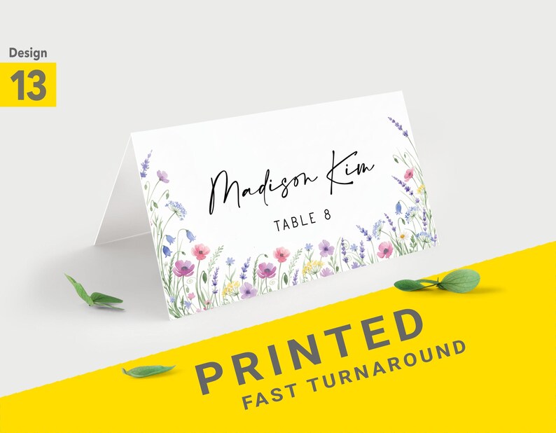 Pu&ograve; includere: Un segnaposto bianco piegato con un design floreale ad acquerello. La carta presenta il nome "Madison Kim" e "Table 8" in corsivo nero. L'angolo in basso a destra riporta il testo "PRINTED FAST TURNAROUND" in grigio.