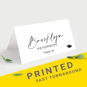 Pu&ograve; includere: Segnaposto bianco piegato con il nome "Brooklyn Peterson" in elegante scrittura nera. La carta include un'icona a forma di foglia e "Table 8". Le parole "PRINTED FAST TURNAROUND" sono stampate su sfondo giallo.