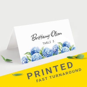 Pu&ograve; includere: Segnaposto piegato bianco con design floreale di ortensie blu. La carta presenta il nome "Brittany Olson" e "TABLE 3" in corsivo nero. Le parole "PRINTED FAST TURNAROUND" sono stampate su sfondo giallo.