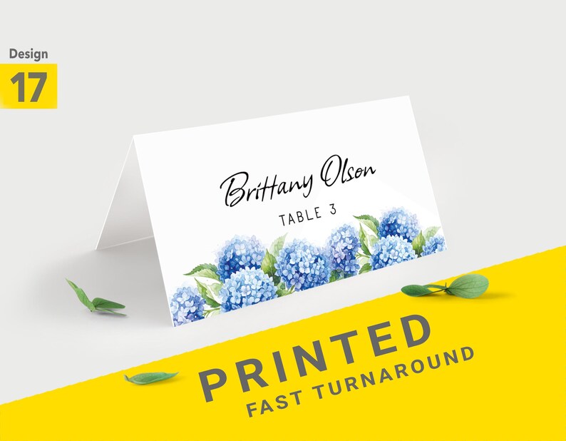 Pu&ograve; includere: Segnaposto piegato bianco con design floreale di ortensie blu. La carta presenta il nome "Brittany Olson" e "TABLE 3" in corsivo nero. Le parole "PRINTED FAST TURNAROUND" sono stampate su sfondo giallo.