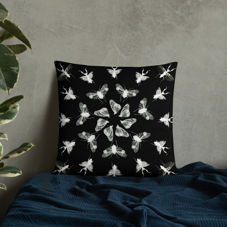 K&ouml;nnte beinhalten: Schwarzes und wei&szlig;es Kissen mit einem kreisf&ouml;rmigen Design aus Insekten und Motten. Das Kissen liegt auf einer blauen Decke.