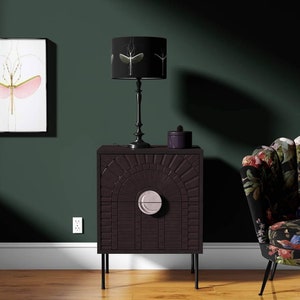 Op de afbeelding: Een donkerbruin nachtkastje met een zonnesterontwerp en een zilveren handvat. Een zwarte lamp met een patroonlampenkap staat erop, naast een kleine paarse doos. De kamer heeft een donkergroene muur en een bloemen fauteuil.