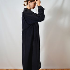May include: A woman wearing a long, dark blue coat with a button-up front. She is standing on a wooden floor with a white wall behind her.