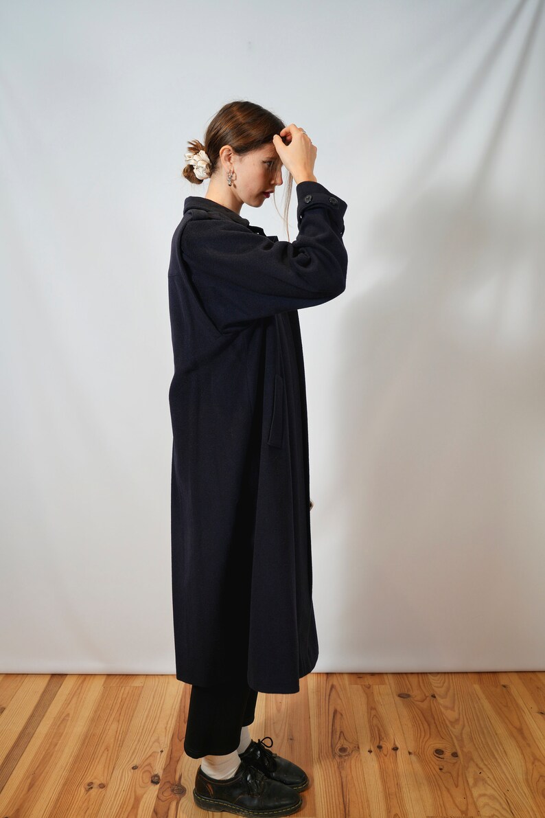 May include: A woman wearing a long, dark blue coat with a button-up front. She is standing on a wooden floor with a white wall behind her.