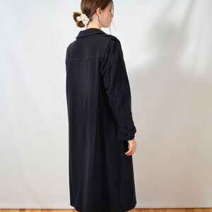 May include: A long, dark blue coat with a collar and long sleeves. The coat is worn by a person with their back to the camera, standing on a wooden floor.