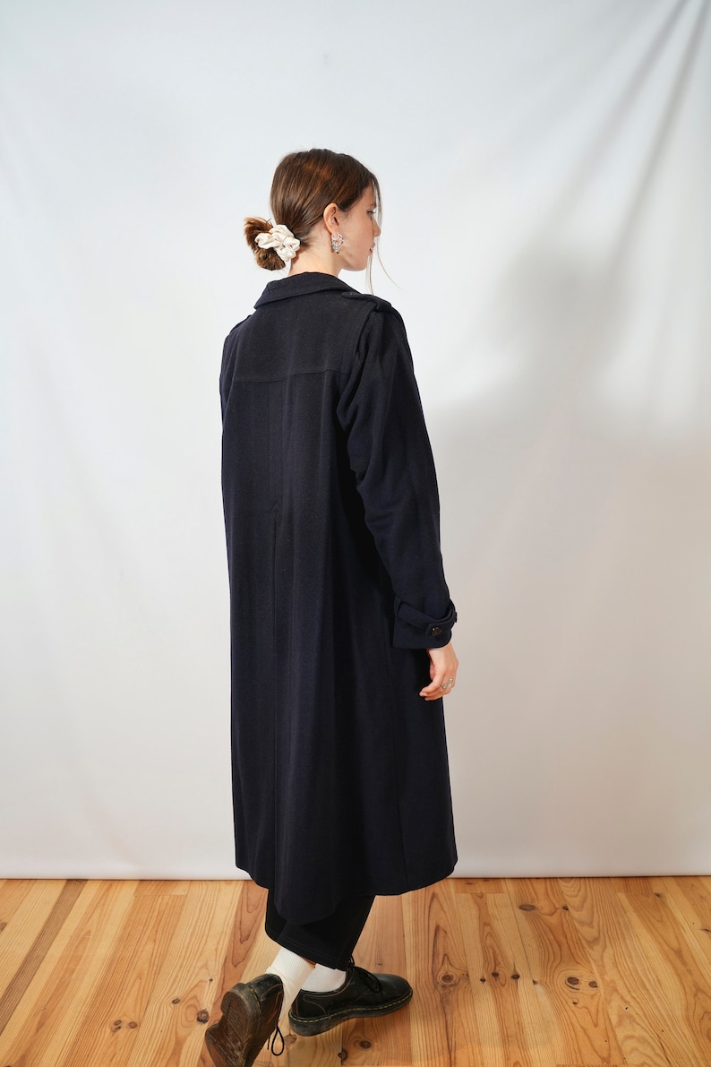 May include: A long, dark blue coat with a collar and long sleeves. The coat is worn by a person with their back to the camera, standing on a wooden floor.