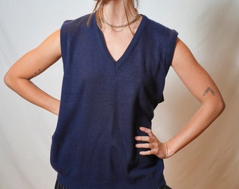May include: A navy blue sleeveless sweater vest with a v-neck. The vest is worn over a black and white striped skirt.