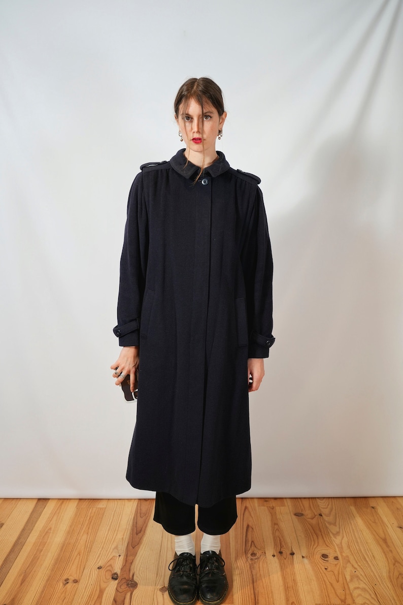 May include: A long, dark blue coat with a button closure and pockets. The coat is worn by a person with dark hair and red lipstick.