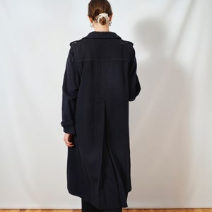 May include: A long, dark blue coat with a single-breasted closure and a long, flowing silhouette. The coat is worn by a person with their back to the camera, standing on a wooden floor.