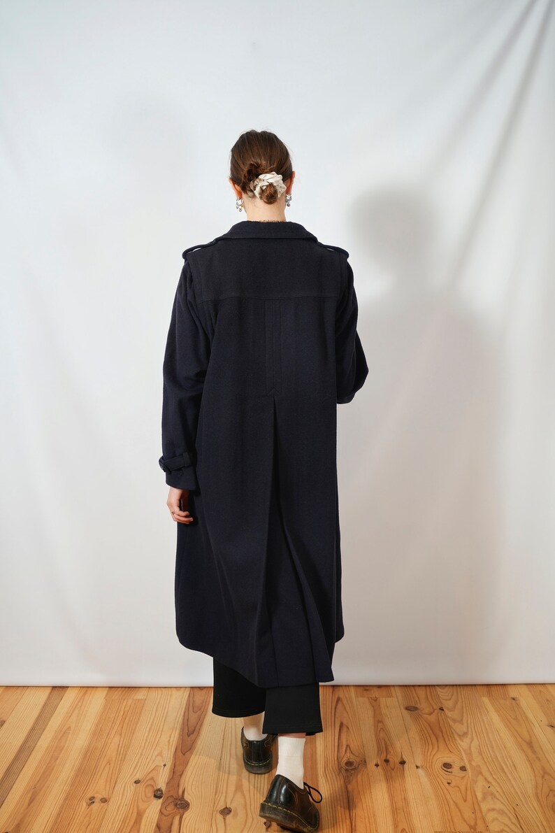 May include: A long, dark blue coat with a single-breasted closure and a long, flowing silhouette. The coat is worn by a person with their back to the camera, standing on a wooden floor.