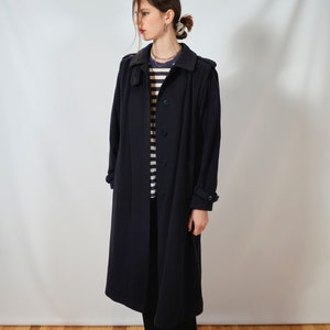 May include: A long, dark blue coat with a button closure and a collar. The coat is worn by a person with a striped shirt and black pants.