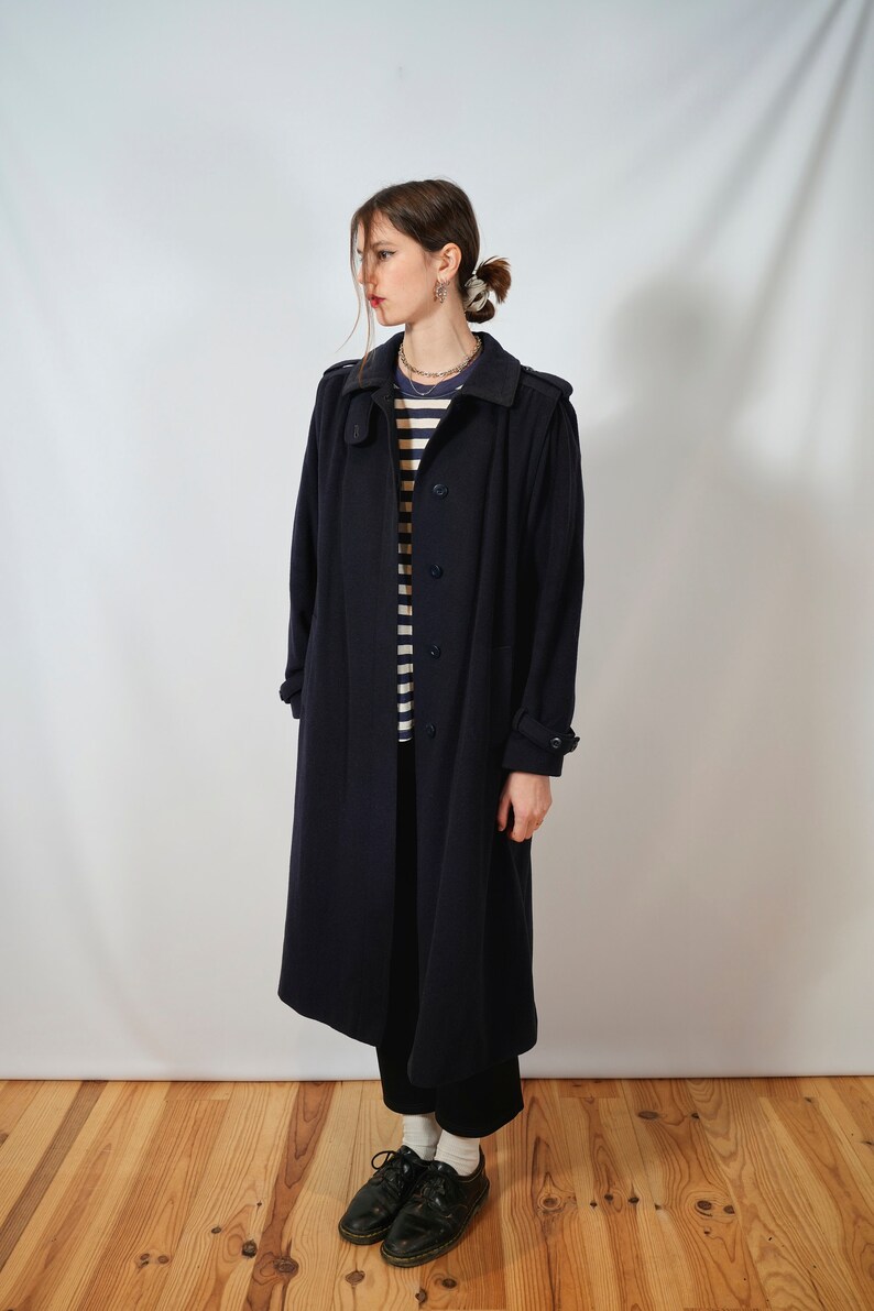 May include: A long, dark blue coat with a button closure and a collar. The coat is worn by a person with a striped shirt and black pants.
