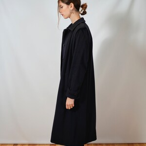 May include: A long, dark blue coat with a double-breasted closure and a collar. The coat has a slightly fitted silhouette and is worn by a person with dark hair.