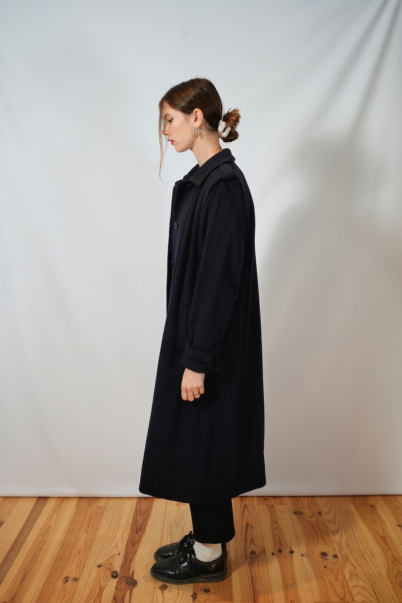 May include: A long, dark blue coat with a double-breasted closure and a collar. The coat has a slightly fitted silhouette and is worn by a person with dark hair.