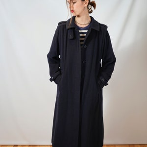 May include: A long, navy blue coat with a collar and pockets. The coat is worn by a person with a striped shirt and a necklace.