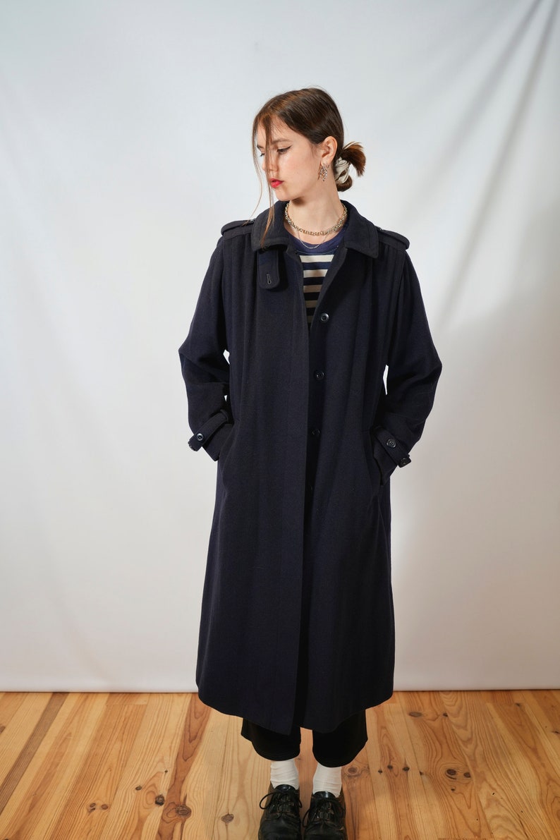 May include: A long, navy blue coat with a collar and pockets. The coat is worn by a person with a striped shirt and a necklace.