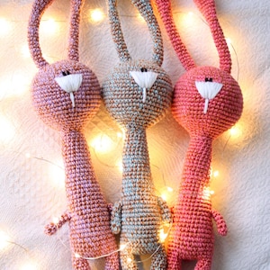 May include: Three handmade crochet bunny dolls in shades of pink and blue. Each bunny has long ears, a white nose, and small arms and legs. The bunnies are arranged on a white surface with string lights.