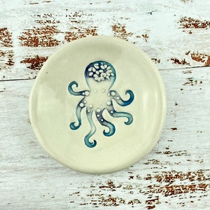 Peut inclure: Un petit plat en céramique blanche avec un motif d'octopus bleu et blanc. La pieuvre a un corps blanc avec des tentacules bleus et des points blancs.