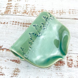 May include: A handmade ceramic soap dish in a soft, seafoam green. The dish has an unusual, asymmetrical shape with a rippled edge. The surface features a decorative pattern of blue and white crystalline formations.