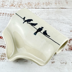 Peut inclure: Un porte-savon en céramique de couleur crème avec une finition brillante, orné d'un dessin au trait noir représentant quatre oiseaux perchés sur un fil. Le porte-savon présente une forme incurvée et ergonomique pour le savon.