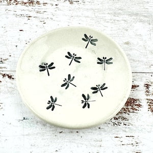 May include: A small, off-white ceramic dish with a slightly raised edge. The dish is decorated with several black dragonfly illustrations. The background is a white, weathered wooden surface.