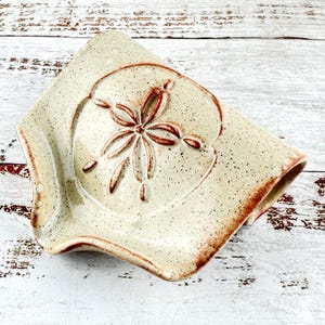 May include: A cream-coloured ceramic spoon rest with a speckled finish and a reddish-brown sand dollar design embossed in the centre. The piece features a curved, concave shape with raised edges and a smooth, glazed surface throughout the entire object.